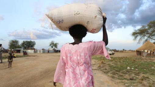 Une femme sud-soudanaise en robe rose s'éloigne de la caméra le long d'un chemin de terre, portant un sac de lentilles et de céréales sur la tête.