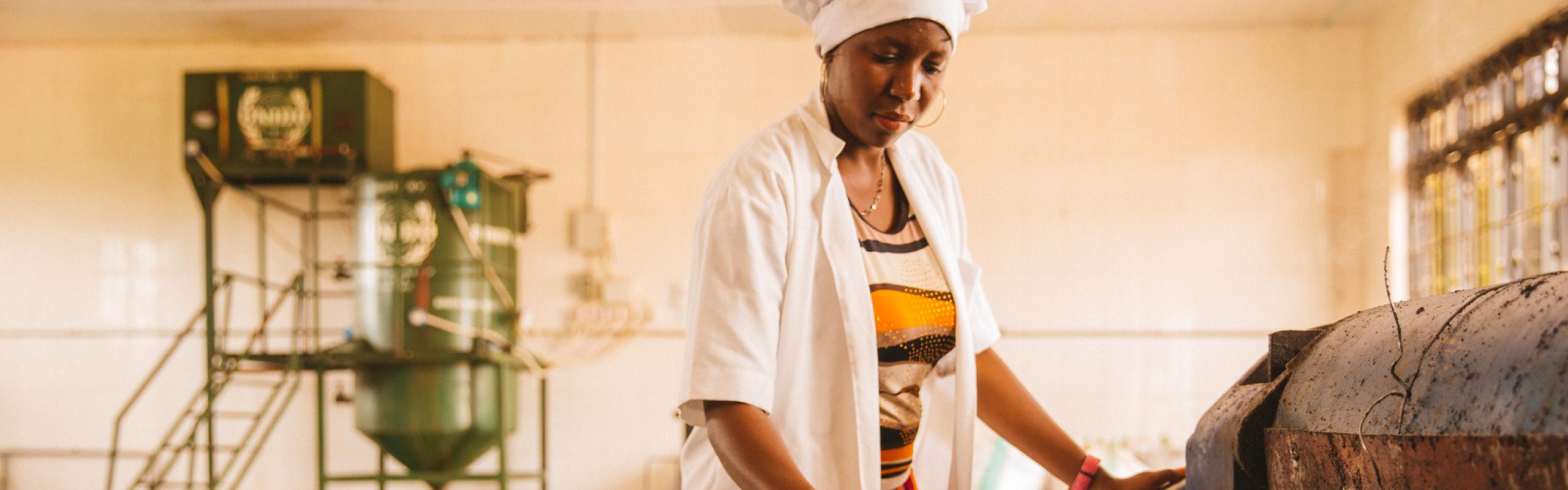 Factory worker in white hat, white jacket, and colorful dress scrapes seed pulp from extraction machine in Dodoma, Tanzania.