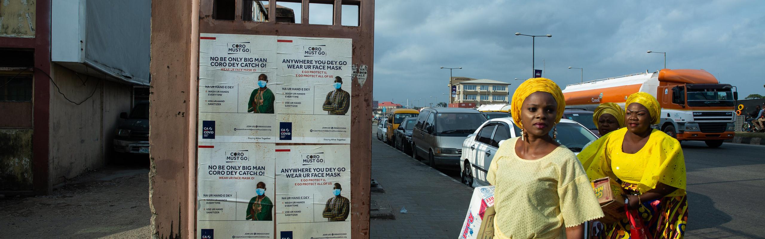 Two women in Lagos, Nigeria carry food and supplies past signs about COVID-19