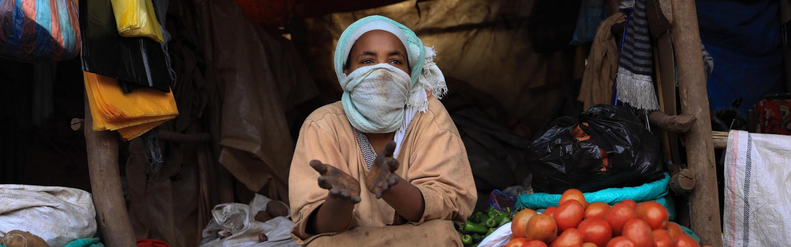 ne femme portant un masque facial en tissu est assise sur le marché de Saris en Éthiopie, entourée de sacs en tissu et de tas d'oignons, de pommes de terre et de tomates.