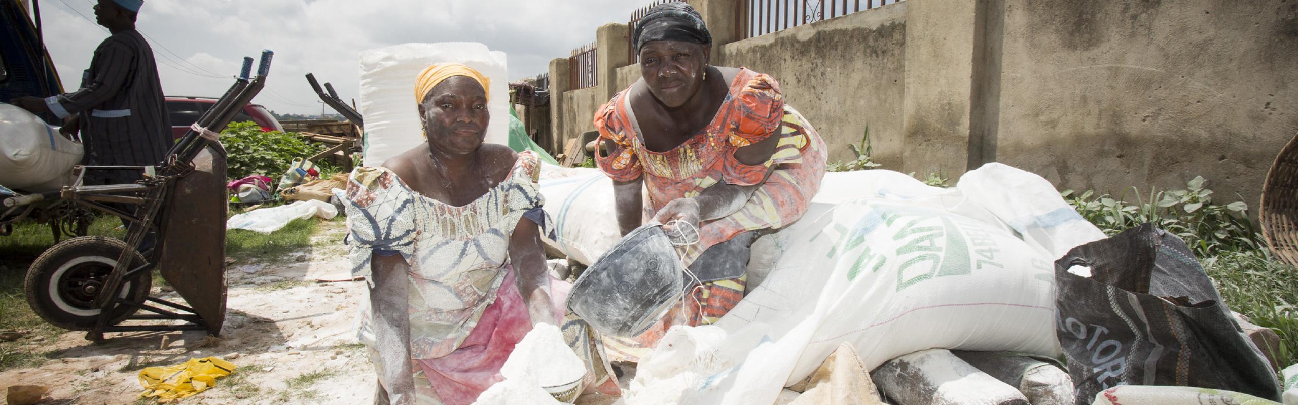 Two women crouch in open air market, sifting cassava flour out of sacks into large bowl
