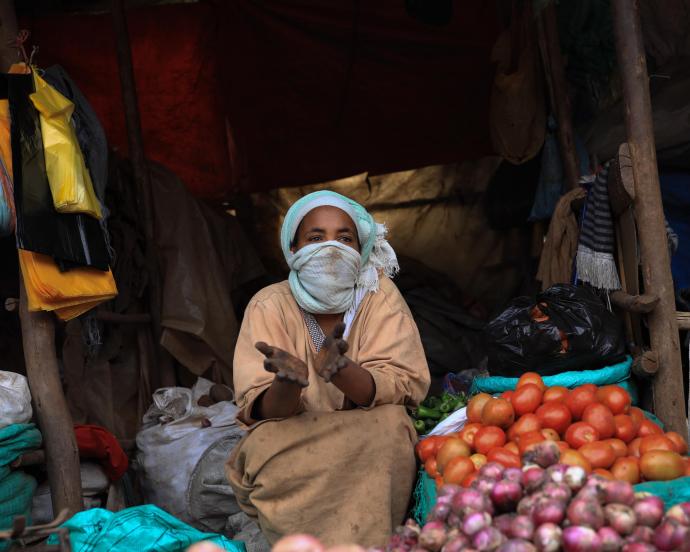 ne femme portant un masque facial en tissu est assise sur le marché de Saris en Éthiopie, entourée de sacs en tissu et de tas d'oignons, de pommes de terre et de tomates.