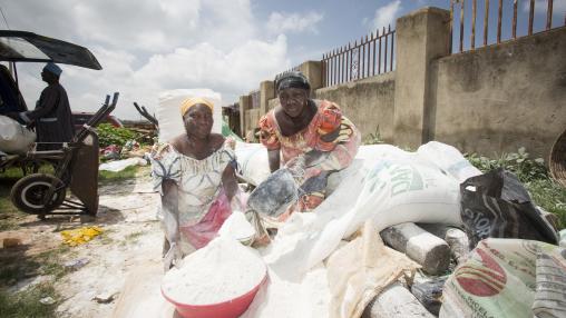 Two women crouch in open air market, sifting cassava flour out of sacks into large bowl