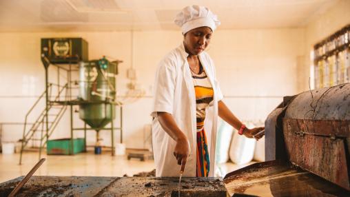 Factory worker in white hat, white jacket, and colorful dress scrapes seed pulp from extraction machine in Dodoma, Tanzania.