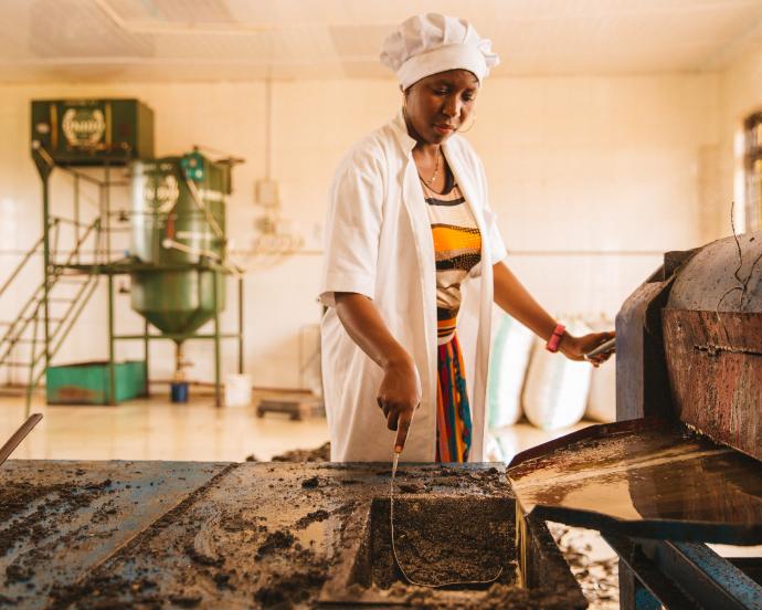 Factory worker in white hat, white jacket, and colorful dress scrapes seed pulp from extraction machine in Dodoma, Tanzania.